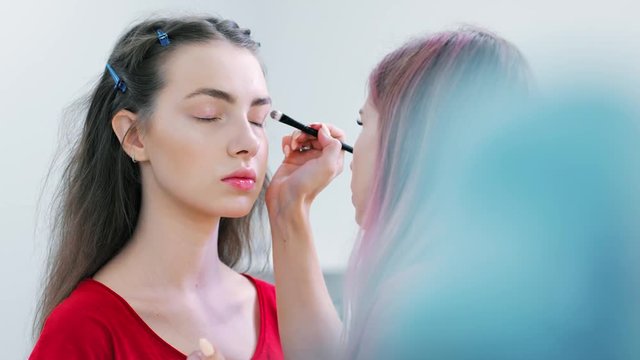 Professional make-up artist putting light primer for eyelid of young European woman client