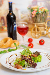 Salad with the fried lard, leaves of lettuce, corn, onions. There are bottle of wine, glass with wine, doughnuts, tomatoes and vase with flowers in the background.