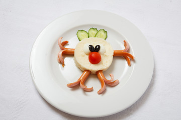 Figure of bob on a white dish. Round trunk from the mashed potatoes, hands and feet from sausages, hair-do from the slices of cucumber, nose from a tomato, eye from olives.