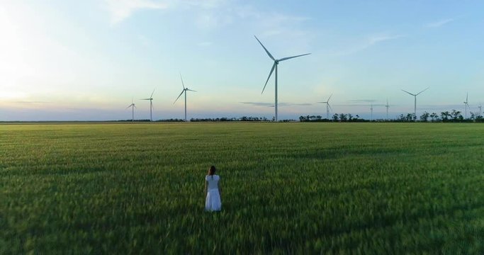 Beautiful girl standing on a green wheat field with windmills for electric power production