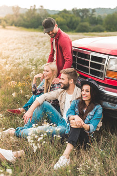 Group Of Young Car Travellers Sitting On Flower Field And Leaning Back On Vintage Truck