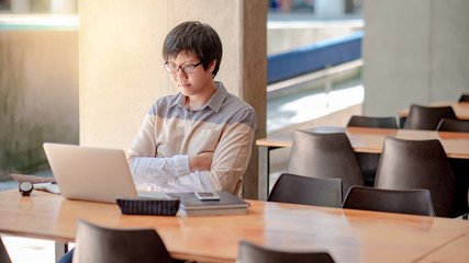 Young Asian man with eyeglasses working with laptop computer sitting in college building. Male university student doing research and self studying. Education and technology concepts