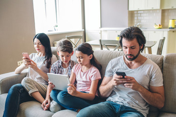 Another picture of family sitting together. Parents hold hpones in hands and look at it. Girl has tablet. She and her brother look at it.