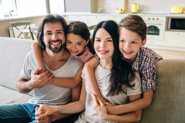 Amazing and beautiful family are sitting on sofa. Children are hugging their parents behind. All of them look on camera and smile.