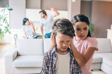 Girl stands behind her brother and keeps his ears closed with hands. Boy is terrified. He has eyes...