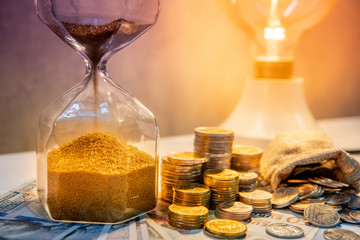 Sand running through the shape of hourglass on table with banknotes and coin stack of international...