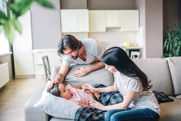 Sick girl is lying on couch. She covered with blanket. Woman holds thermometer and look at it. Guy...