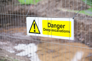 Deep excavations danger sign on fence with trench hole in background on construction site