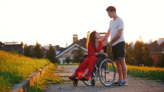 Girl In A Wheelchair Cute Talking To Her Young Man On The Background Of The House, Green Grass And Trees
