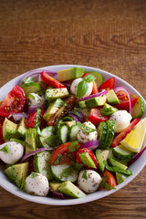 Avocado, tomato, cucumber salad with mozzarella cheese in white bowl on wooden table. vertical