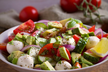 Avocado, tomato, cucumber salad with mozzarella cheese in white bowl on wooden table. horizontal