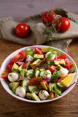 Avocado, tomato, cucumber salad with mozzarella cheese in white bowl on wooden table. vertical