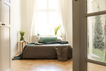 Green blanket on grey bed in minimal bedroom interior with flowers and window. Real photo