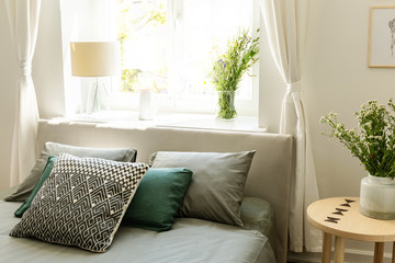 Patterned green and grey pillows on bed next to table with flowers in bedroom interior. Real photo