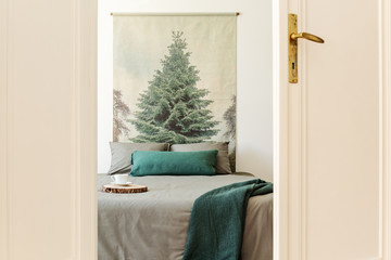 Green blanket on grey bed against tree painting in white simple bedroom interior. Real photo