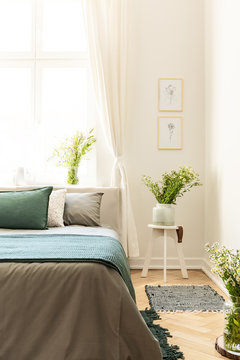 Posters on white wall above flowers on table next to grey and green bed in bedroom interior. Real photo