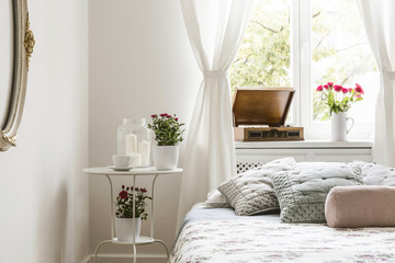 White bedroom interior in real photo with bedside table with tea cup, candles and red roses,...