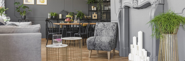 Real photo of an armchair standing next to small tables and a couch in a living room interior with table with black chairs and shelves with ornaments in the background