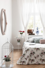 Real photo of bright bedroom interior with window with curtains, fresh roses in bird cage, bed with floral bedding and herringbone parquet