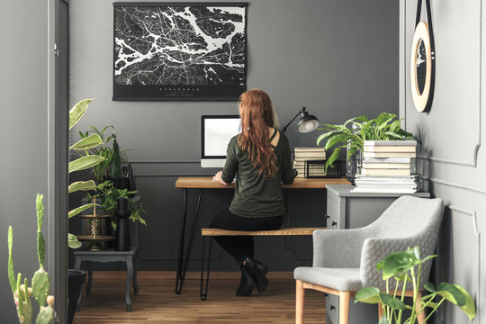 Ginger haired woman sitting by the hairpin wooden desk with mockup computer in real photo of dark grey living room interior
