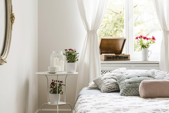 White bedroom interior in real photo with bedside table with tea cup, candles and red roses, windowsill with gramophone and fresh flowers