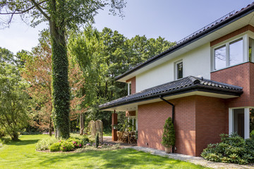 Red brick wall of house with roof and windows next to trees and garden