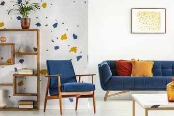 Armchair and couch with pillows in blue and orange living room interior with poster and plant. Real...