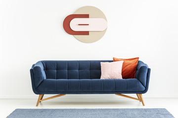 Pink and orange pillows on blue sofa in white apartment interior with poster and carpet. Real photo
