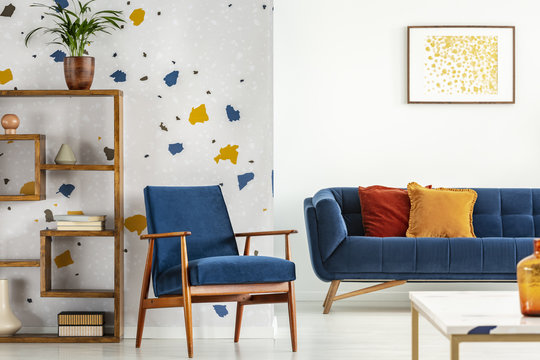 Armchair and couch with pillows in blue and orange living room interior with poster and plant. Real photo