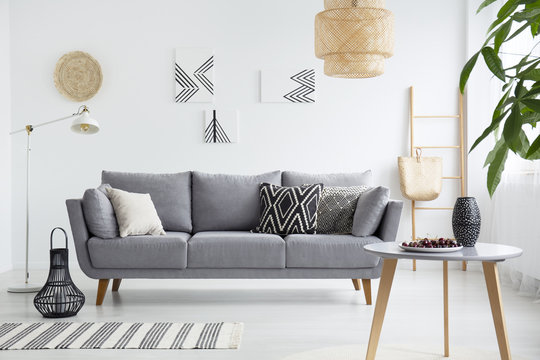 Real photo of a scandi living room interior with cushions on gray couch, cherries on wooden table and bag on a ladder