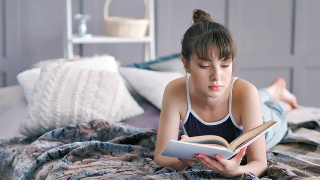 Full shot attractive brunette girl lying on stomach crossing bare feet on bed and reading book