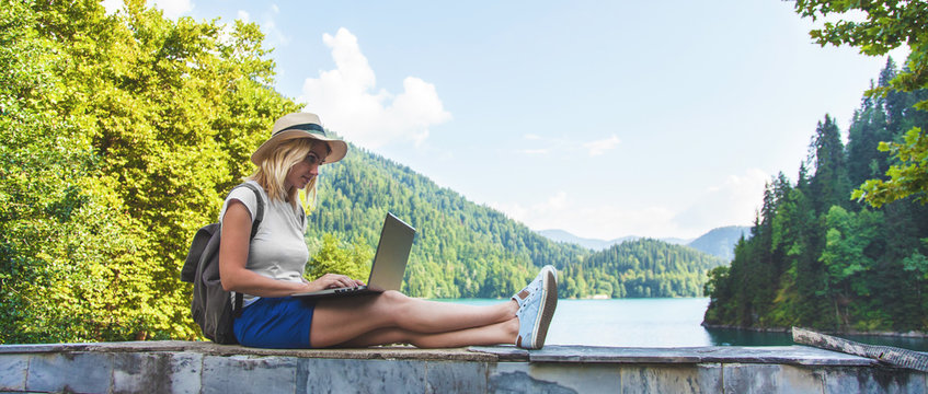 Beautiful girl traveler in a hat on a lake with a laptop Work outside the office