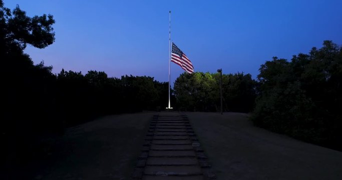 Drone view of US flag at half-staff at dawn, lit by spotlight,, moving forward and up