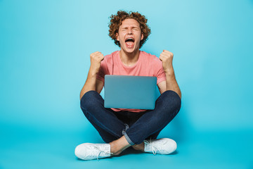 Image of pleased joyous man 20s with brown curly hair rejoicing and clenching fists, while sitting...
