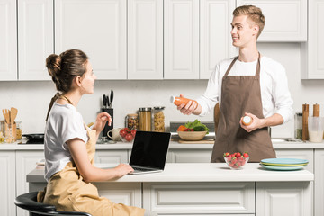 girlfriend using laptop, boyfriend holding bottles of juice in kitchen