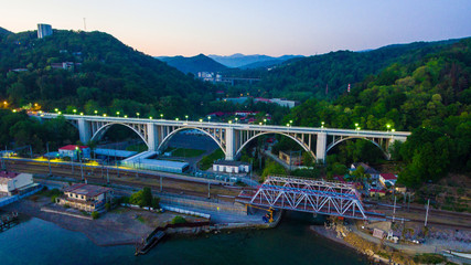 Drone view of the illuminated Matsesta viaduct and railway bridge on the background of mountainsides with dense forest at twilight, Sochi, Russia
