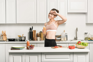 attractive girl in sport bra touching forehead and holding bottle of fresh juice in kitchen