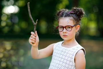 Funny adorable little kid girl with glasses and wooden magic wand playing Harry Potter in park on sunny day. - obrazy, fototapety, plakaty