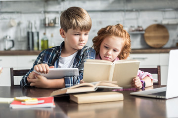 concentrated little brother and sister with devices reading book together