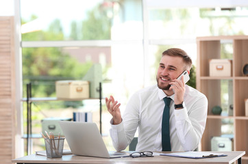 Young businessman talking on phone while using laptop at table in office