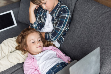 high angle view of little brother and sister relaxing on couch with gadgets