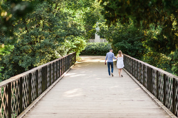 Fototapeta na wymiar Young couple walking along peaceful park scenery on a beautiful summer day. Green canopy of trees and a metal bridge above the water of a pond. Concept of hygge.