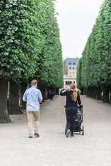 Young parents with a baby relaxing during a summer day in a Danish park, perfectly manicured linden trees line an elegant garden alley. Concept of hygge.