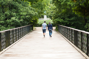 Young couple walking along peaceful park scenery on a beautiful summer day. Green canopy of trees and a metal bridge above the water of a pond. Concept of hygge.