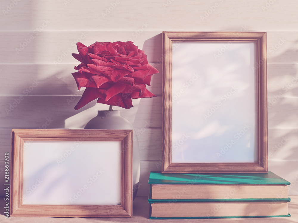 Wall mural A fresh red rose with droplets on the petals is in a gray opaque vase. Nearby there are two empty wooden frames, one of which is on a pile of books. White wooden background