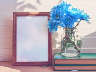 An empty wooden frame is on a white wooden background. Nearby is a glass jar with blue chrysanthemums or daisies on a stack of books.