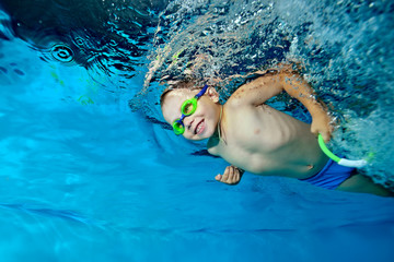 A beautiful little boy is swimming under the water in the pool on a blue background, looking forward and smiling. Portrait. Horizontal orientation © alexbard