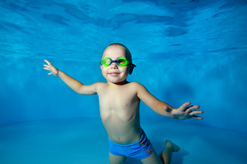The kid is posing under the water at the bottom of the pool on a blue background, looking at me and smiling. Portrait. Horizontal orientation © alexbard