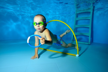 A little boy swims under the water in the pool and collects toys from the bottom. Portrait....
