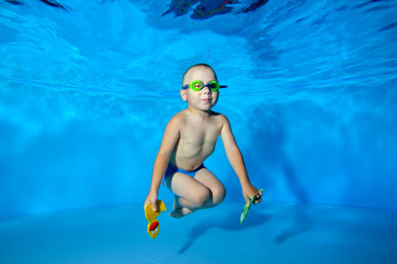 A happy little boy swims and poses underwater in the pool on a blue background with swimming glasses and toys in his hands. He looks at the camera and smiles. Portrait. Underwater photography